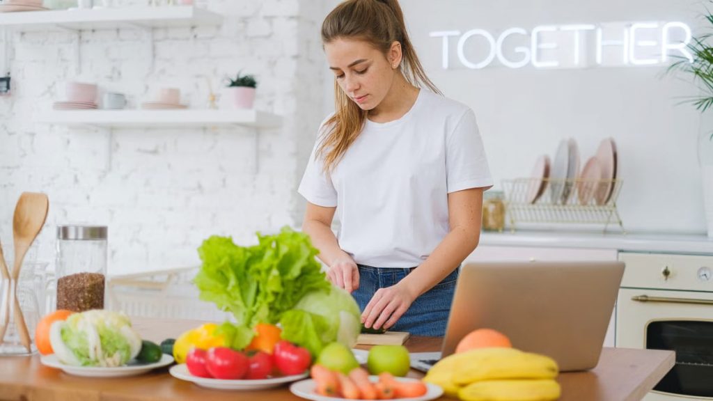 Woman in white t-shirt preparing fresh vegetables in a bright kitchen with a laptop.