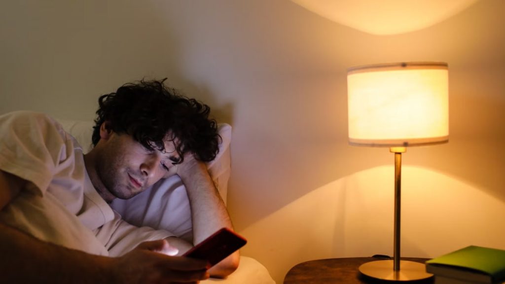 A man lying in bed using a smartphone with a lamp providing warm ambient light.